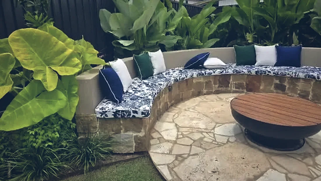 Crazy paved fire pit circle enclosed by a curved sandstone bench seat with cushions and tropical planting