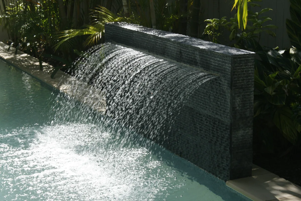 Honed black stone water blade spilling a clear sheet of water into a pool edged with tropical planting