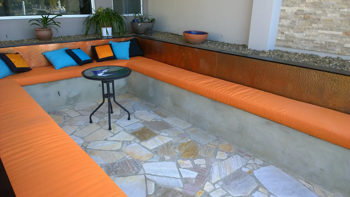 A sunken outdoor lounge floored in sandstone crazy paving, with a built-in wrap-around bench and tangerine cushions