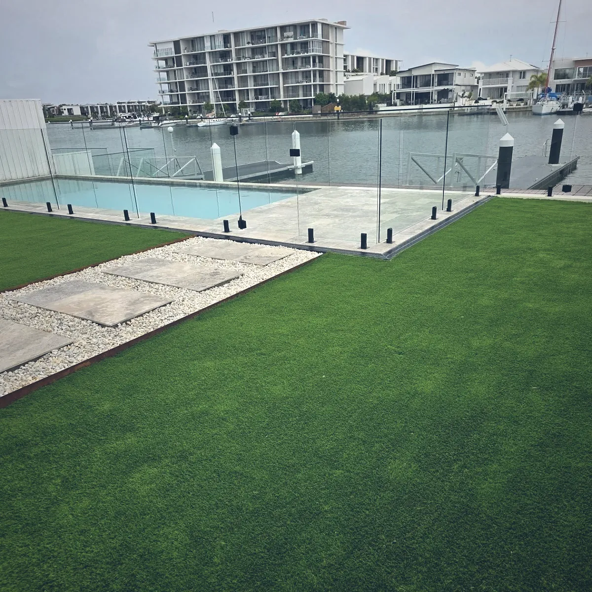 Level waterfront lawn with travertine stepping stones and a glass pool fence